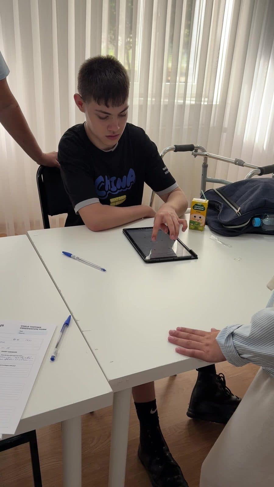 A teenager using a tablet during a structured FlexiKeys usability session, with a mobility walker beside him and a printed Child Testing Observation Form on the table